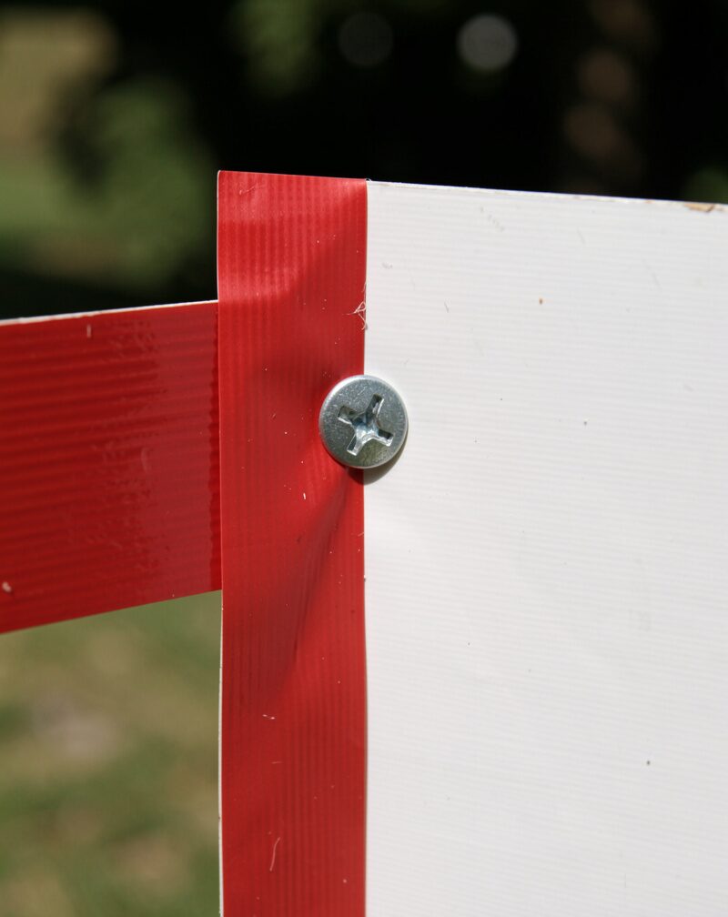 A red corrugated plastic strip lying flat on grass beside a screwdriver and two screws, unprinted side up